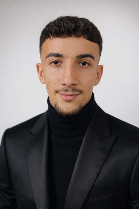 Portrait of a young man with dark hair and facial hair wearing a black blazer and turtleneck against a white background